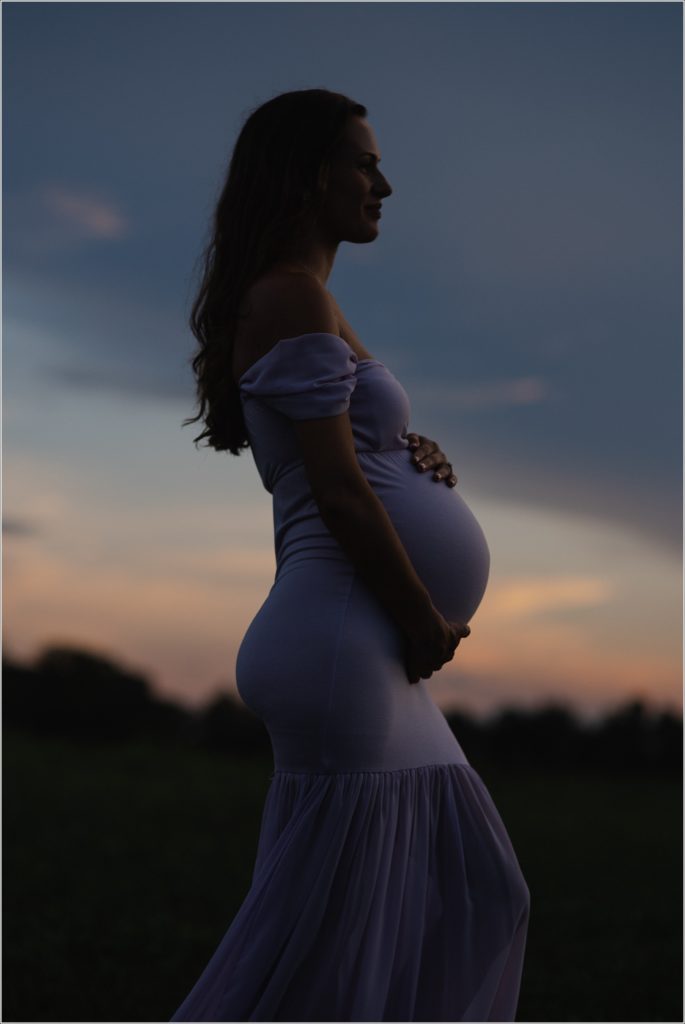 Pregnant woman in lilac maternity dress in Urbana Villages field in sunset silhouette