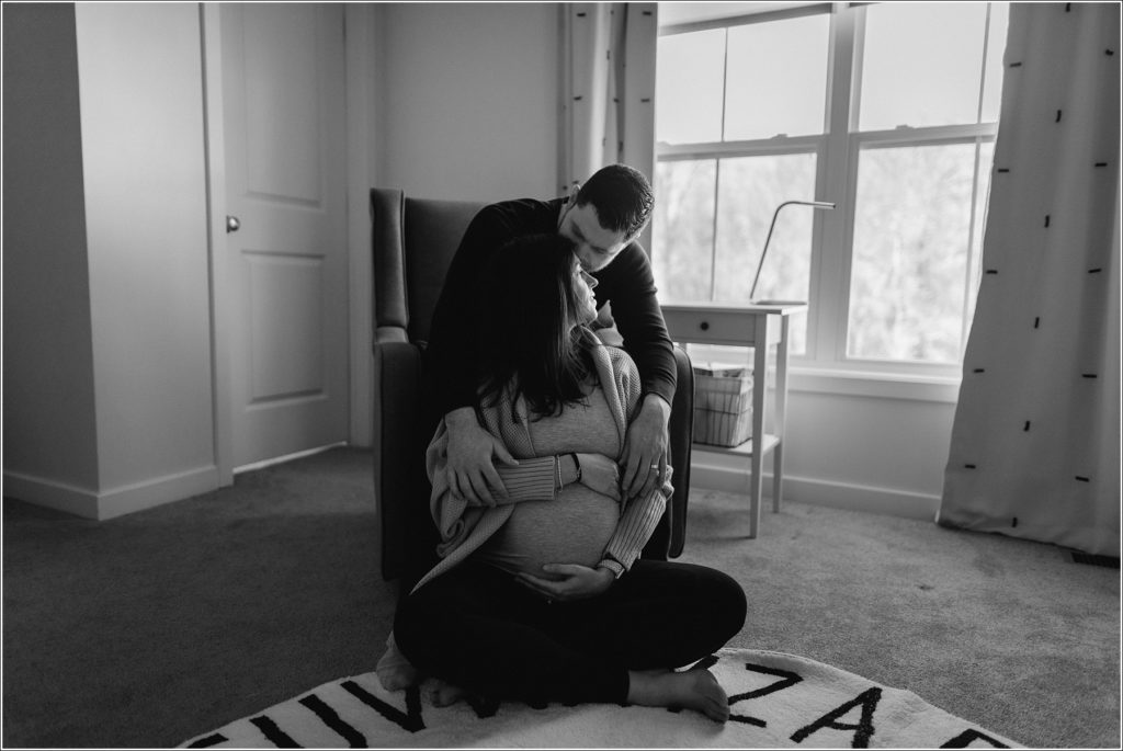 black and white photo couple poses on floor and in chair looking toward window holding pregnant belly
