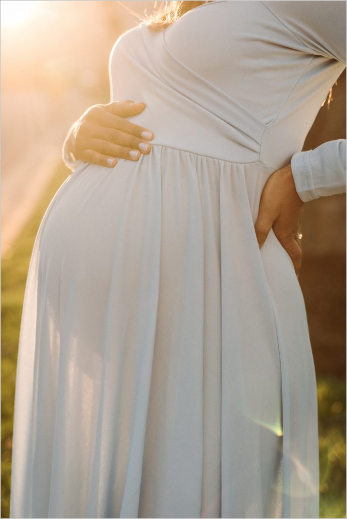 pregnant woman in baby blue maternity dress holds belly at sunset