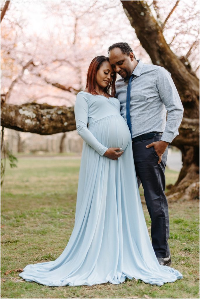 couple poses with pregnant belly in blue maternity dress at sunset in front of kenwood cherry blossoms