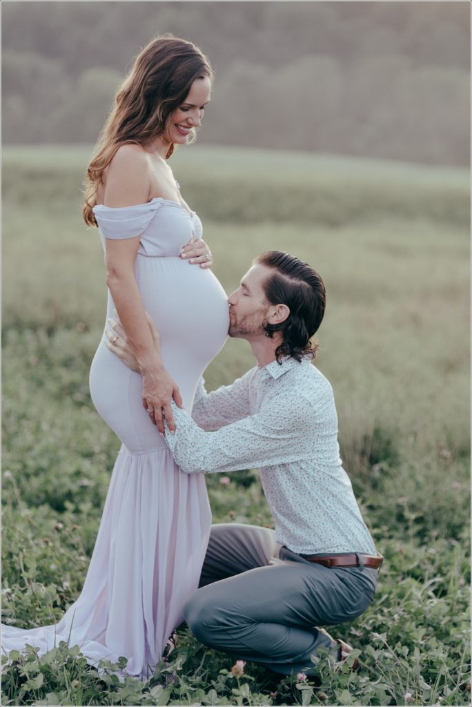 Pregnant woman in lilac maternity dress in Urbana Villages field with her husband and kisses belly