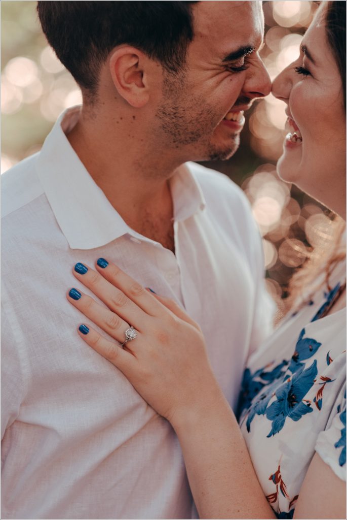 Engaged Couple snuggles by the Georgetown Waterfront in white linen outfits with blue flowers showing off diamond ring