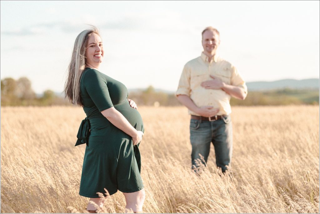 pregnant woman holds belly in field while husband holds belly behind her