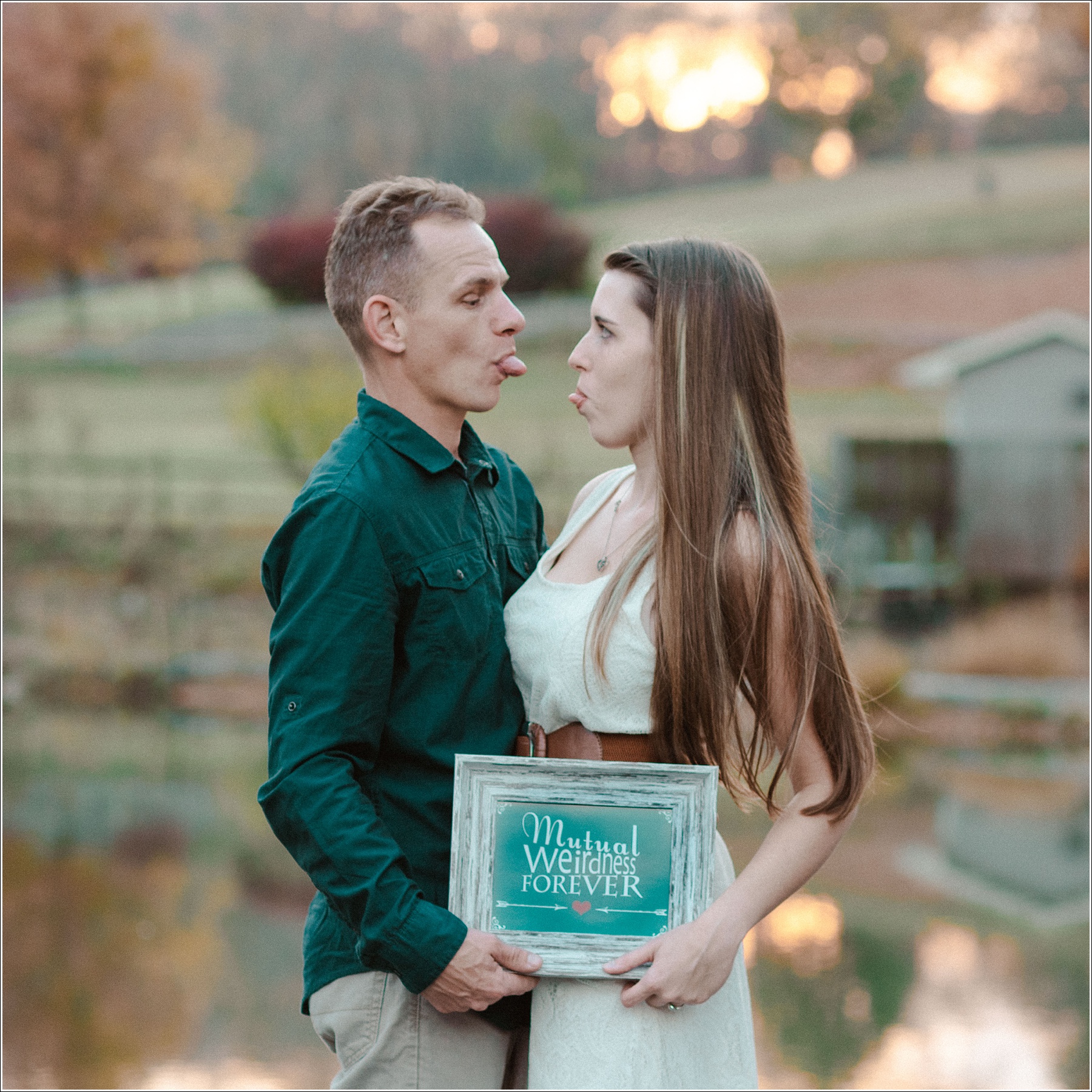 engaged couple sticks tongues out at each other while holding sign that says mutual weirdness forever at steele creek park