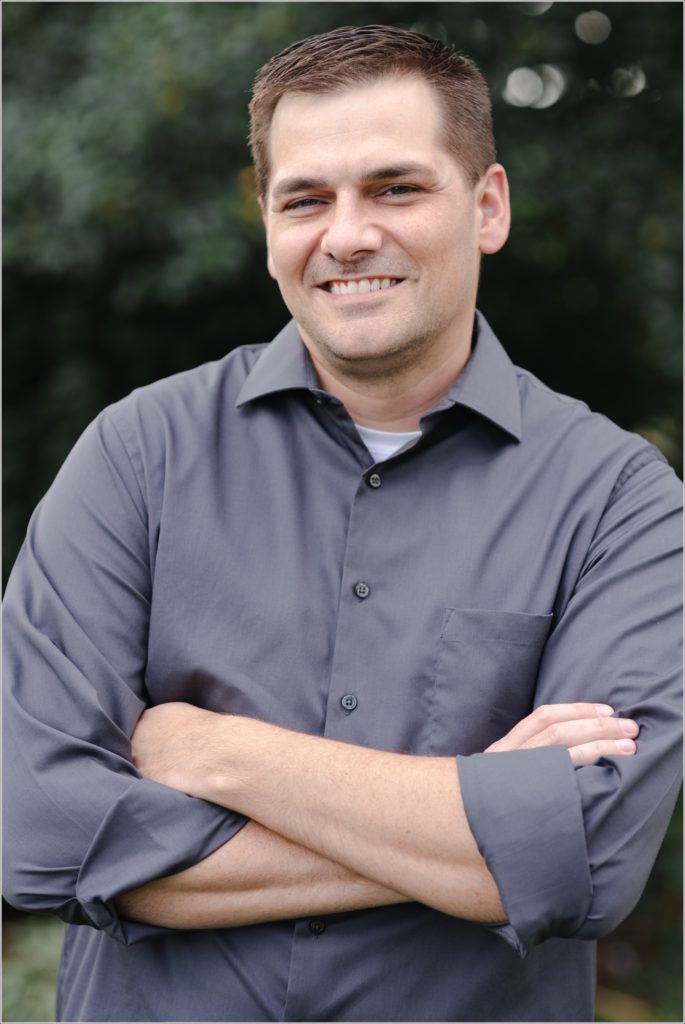 man in grey button up with arms folded smiles for the camera against green backdrop