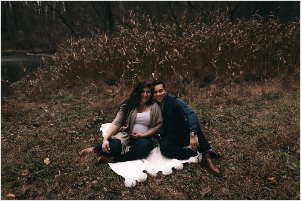pregnant woman and man sit on a blanket in front of a field