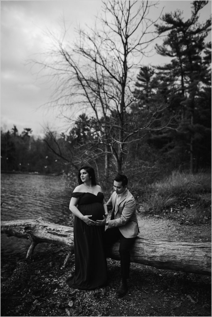 pregnant woman and man sit on a log looking over a lake in black and white