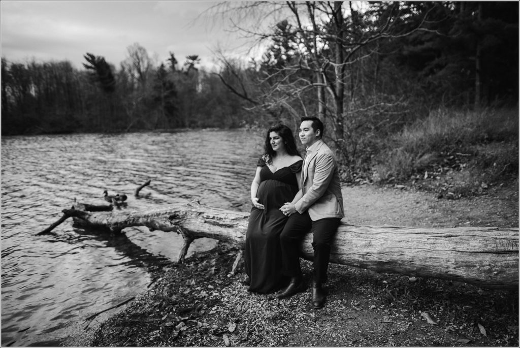 pregnant woman and man sit on a log looking out over a lake black and white photo