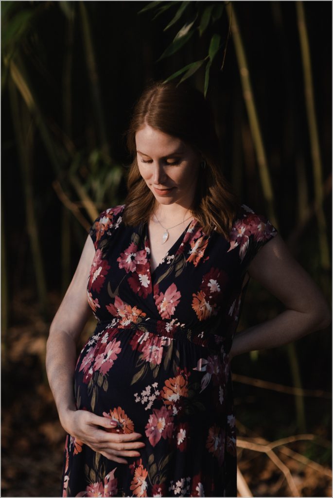 pregnant woman holds her belly wearing a floral print dress with sunset lighting on her face