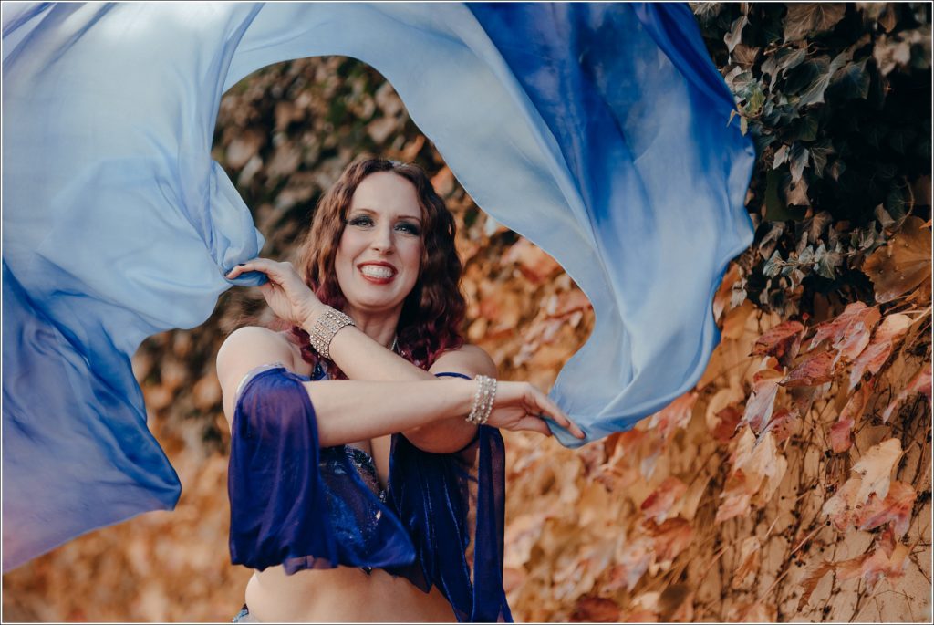belly dancer photoshoot in blue and purple costume in front of wall of orange and green leaves