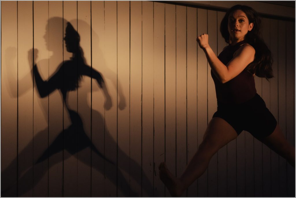 dc dancer jumps into yellow light against white barn with shadows behind her