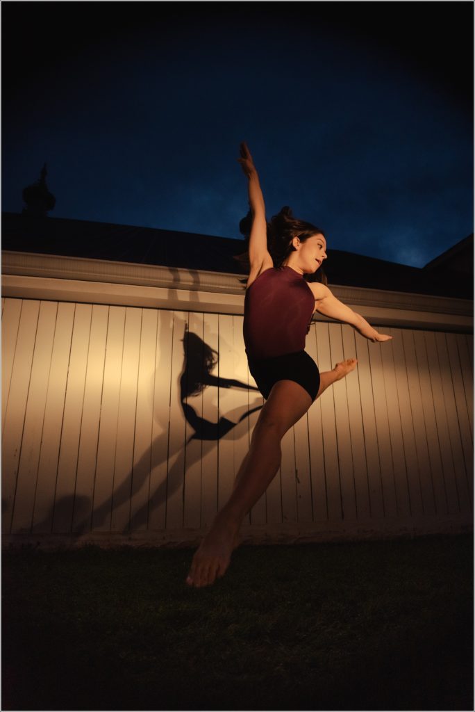 dc dancer jumps into yellow light against white barn with shadows behind her