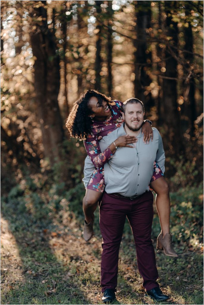 woman in floral dress piggy back on man laughing in front of woods with sunset streaming through for engagement photographers DC