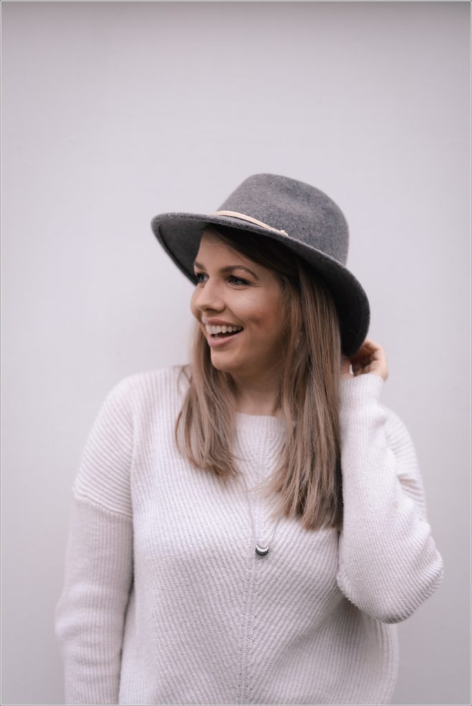 arbonne consultant poses against white wall in white sweater and grey hat for personal branding photography
