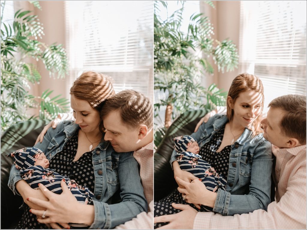 mother and father hold newborn baby by window with green plant in background