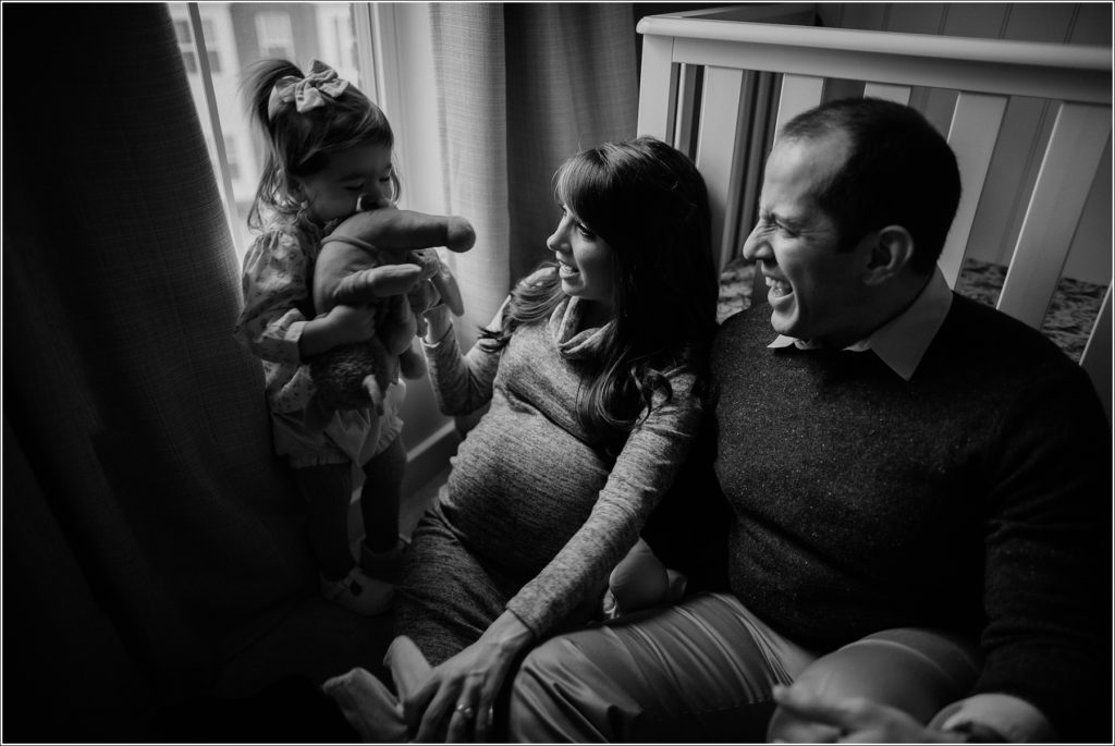 mom and dad and daughter sit near crib in window light for in home maternity photography black and white