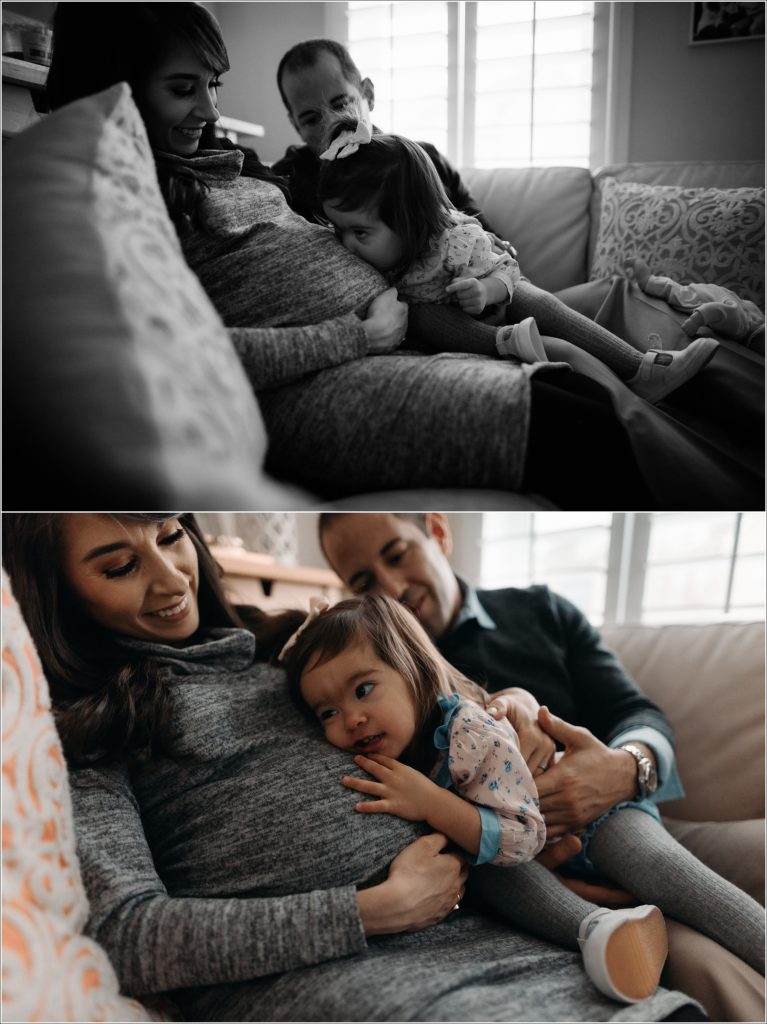 mom and dad and daughter sit on couch and daughter kisses moms belly