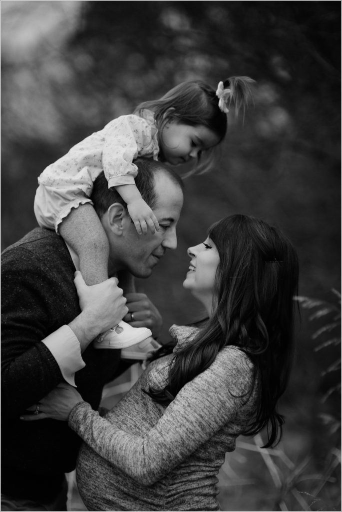 daughter sits on dads shoulders in black and white photo as dad leans in to mom