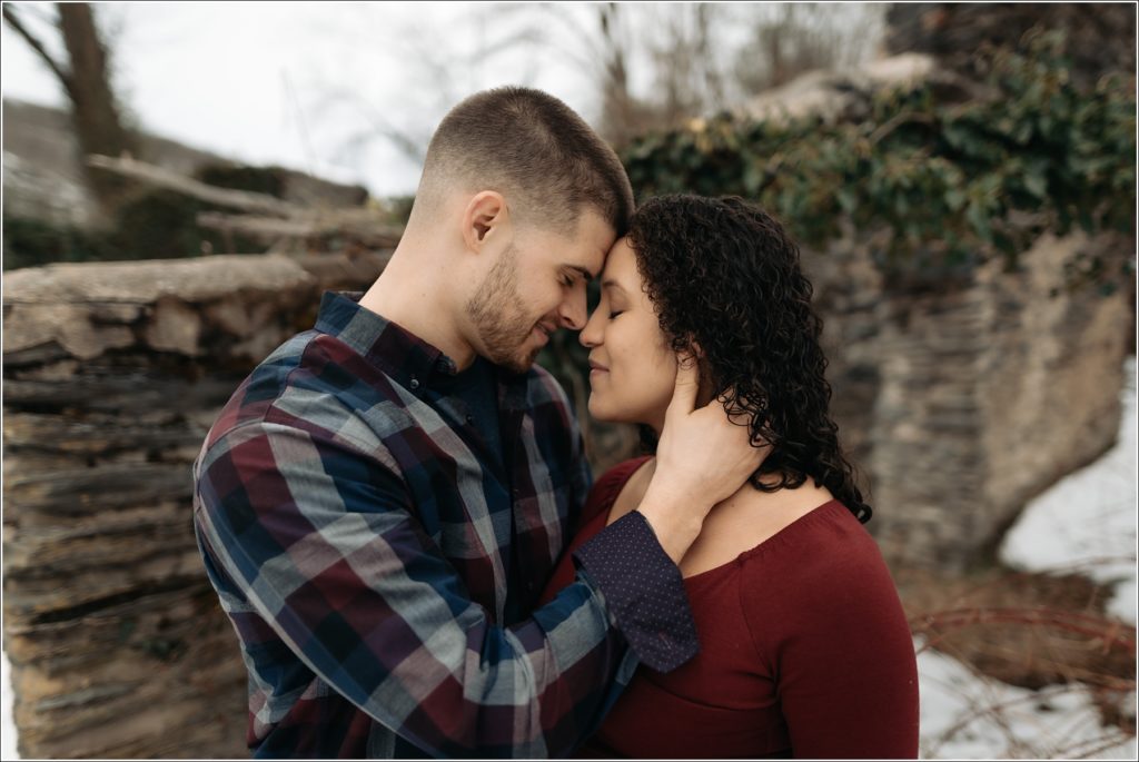 man in maroon and blue plaid shirt and woman in cranberry embrace in front of stone wall