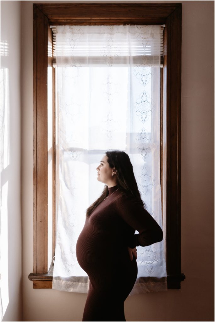 pregnant woman in purple maternity sweater dress poses in photo studio in front of window