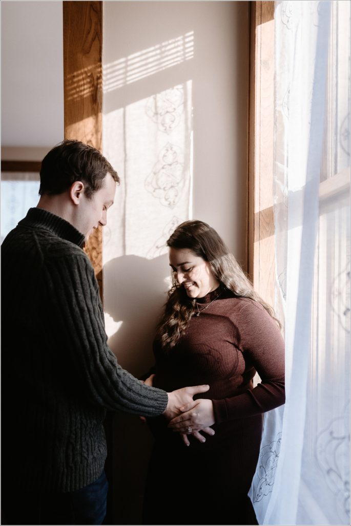 man and woman pose for maternity photos in front of window with lace curtains as sun shines in and creates shadows