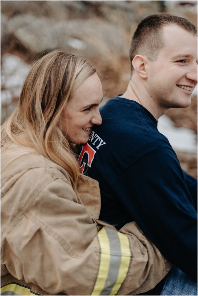 a couple wearing a blue sweater and firefighter hat and coat pose in the snow at middletown overlook for firefighter engagement photos