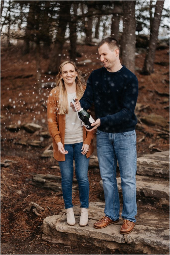 couple in blue and orange sweaters pop champagne at the middletown overlook for firefighter engagement photos