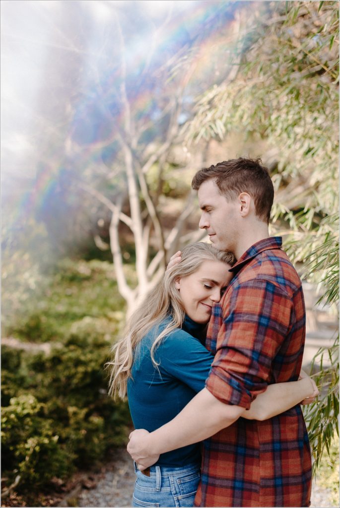 couple poses in asian garden for national arboretum engagement photos in red plaid and a teal turtleneck and jeans with rainbow prism above them hugging