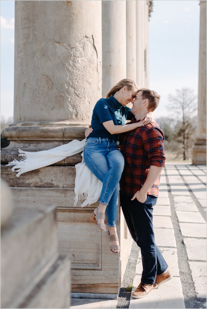 woman sits against columns posing with fiancé for national arboretum engagement photos wearing red plaid and teal turtleneck and jeans