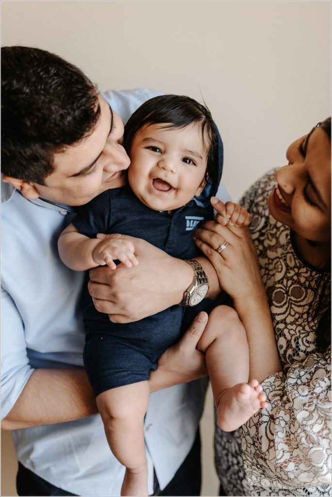 mom and dad pose with smiling baby in dark blue onesie in newborn photography at home