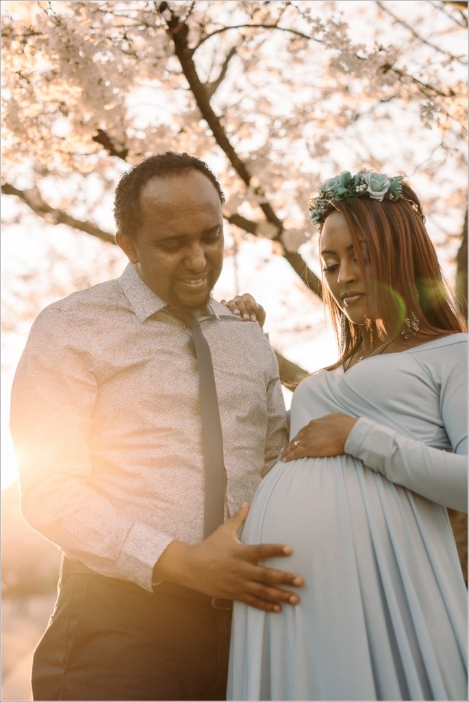 couple poses with pregnant belly in blue maternity dress at sunset in front of kenwood cherry blossoms