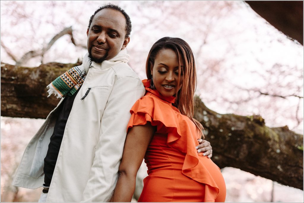 pregnant couple poses in front of kenwood cherry blossoms in white jacket, orange dress and Ethiopian scarf