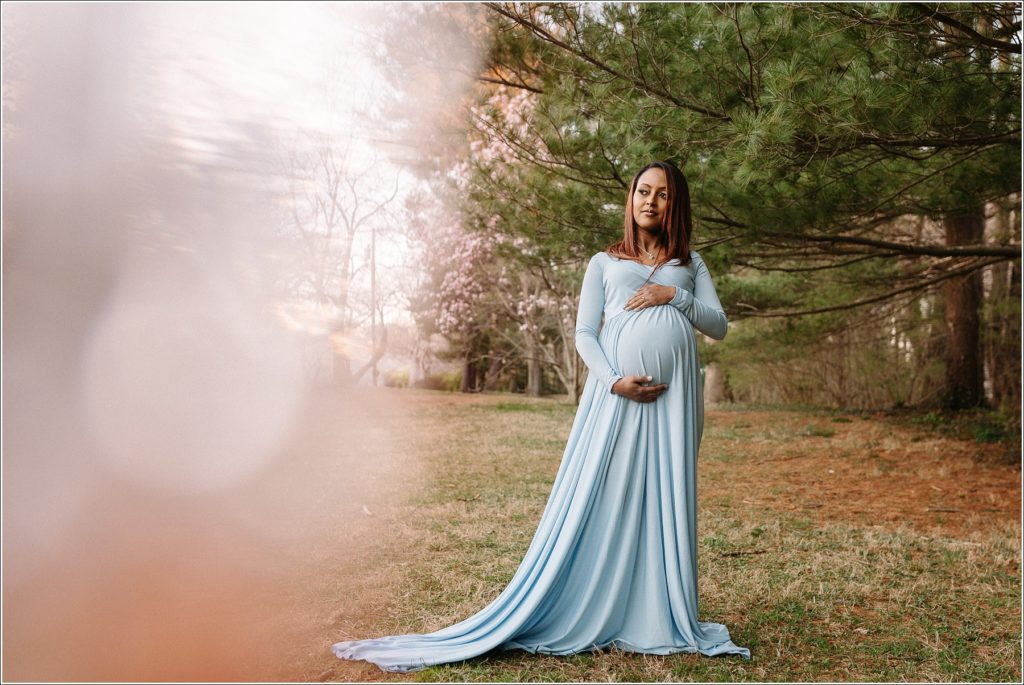 woman in blue maternity dress poses in front of green trees with pink prism