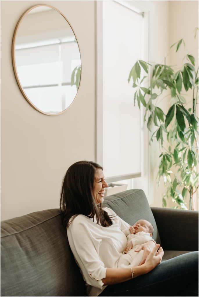 new mother laughs with newborn on couch with plant and mirror behind her