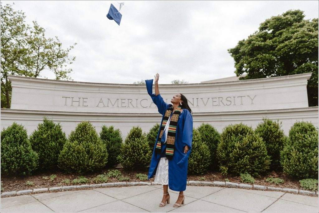 american university graduation photos in front of school in blue cap and gown throwing hat