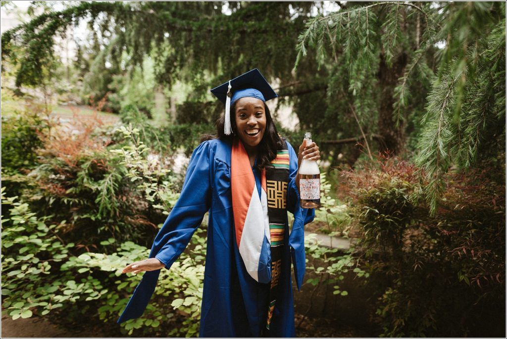 kogod school of business grad in blue cap and gown poses in front of greenery with champagne bottle