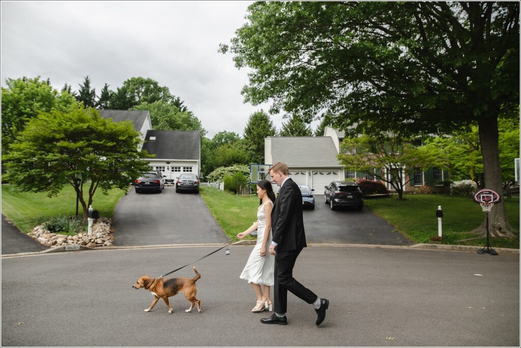 woman in knee length silk wedding dress walks next to man in black suit with dog on leash